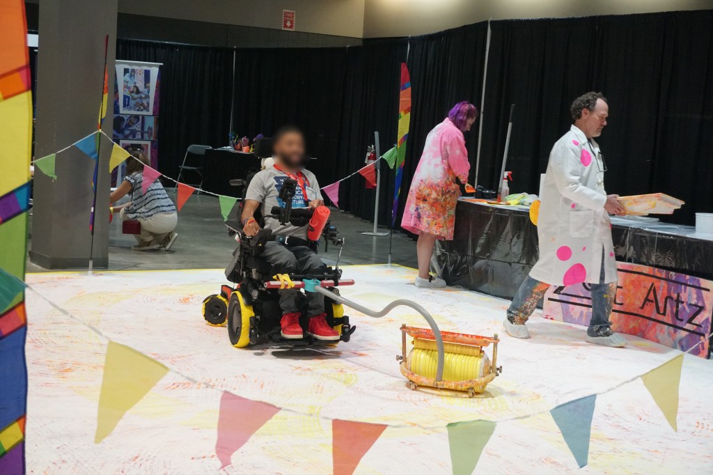 An area sectioned off with colorful flags features a large "canvas" on the floor, with a dark-skinned man in a wheelchair pushing a giant paint roller attached to his chair.