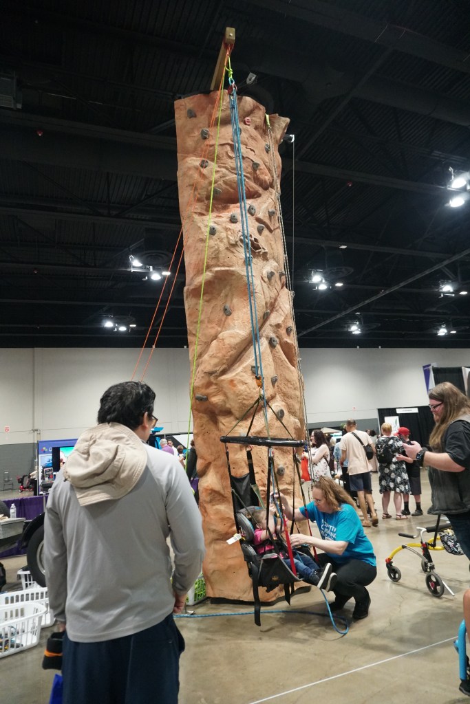 A woman is assisting a small female with the adaptive seat for the climbing wall, with the tall wall poised in the background.