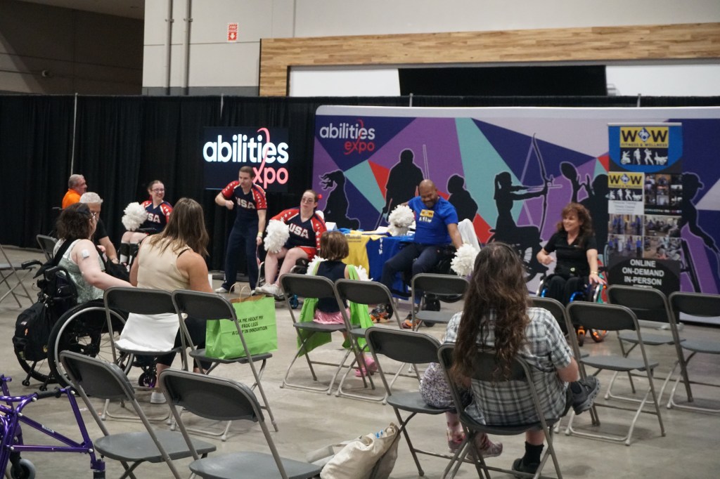 Justin Graham leads a robust workout with pom-poms as several others lead alongside him, with expo attendees joining from their seats.